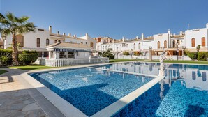 Una piscina con borde de piedra, rodeada de edificios blancos con ventanas arqueadas y una palmera.