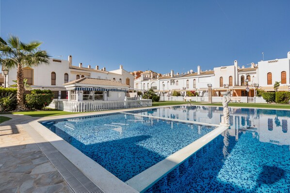 A swimming pool with a stone border, surrounded by white buildings with arched windows and a palm tree.