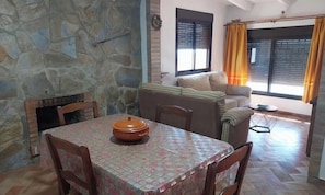 A dining area with a table, chairs, and a large pot. There is a stone wall, a sofa, and a window with curtains.