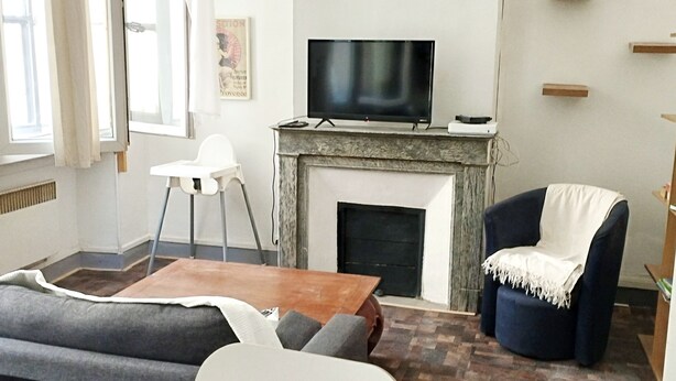 Uma sala de estar com sofá, mesa de centro, televisão sobre a lareira e uma poltrona com cobertor.