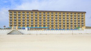 A large hotel with multiple floors and balconies, a covered outdoor area with blue seating, and a sandy beach in the foreground.