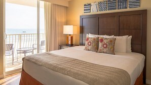 A hotel room with a large bed, a nightstand with a lamp, a view of the ocean, and a balcony with chairs.