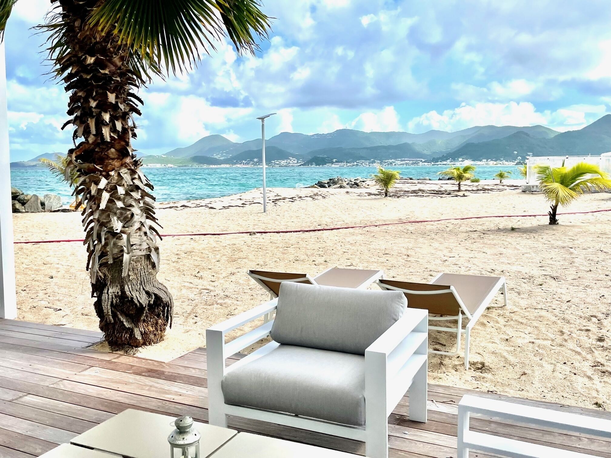A beachside lounge with a palm tree, white cushioned chairs, and a view of the ocean and mountains.