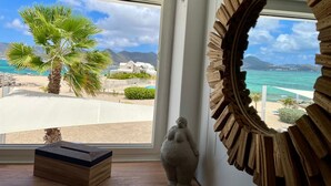 A room with a large window overlooking a beach, a wooden sculpture, and a stone figure. - Beach front fabulous new all renovated unit 3 bedrooms 3 Baths, pools see view (Collectivité de Saint-Martin)