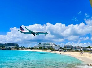 Un avion de la compagnie American Airlines survole une plage bordée d’une eau turquoise cristalline, avec un hôtel en arrière-plan.