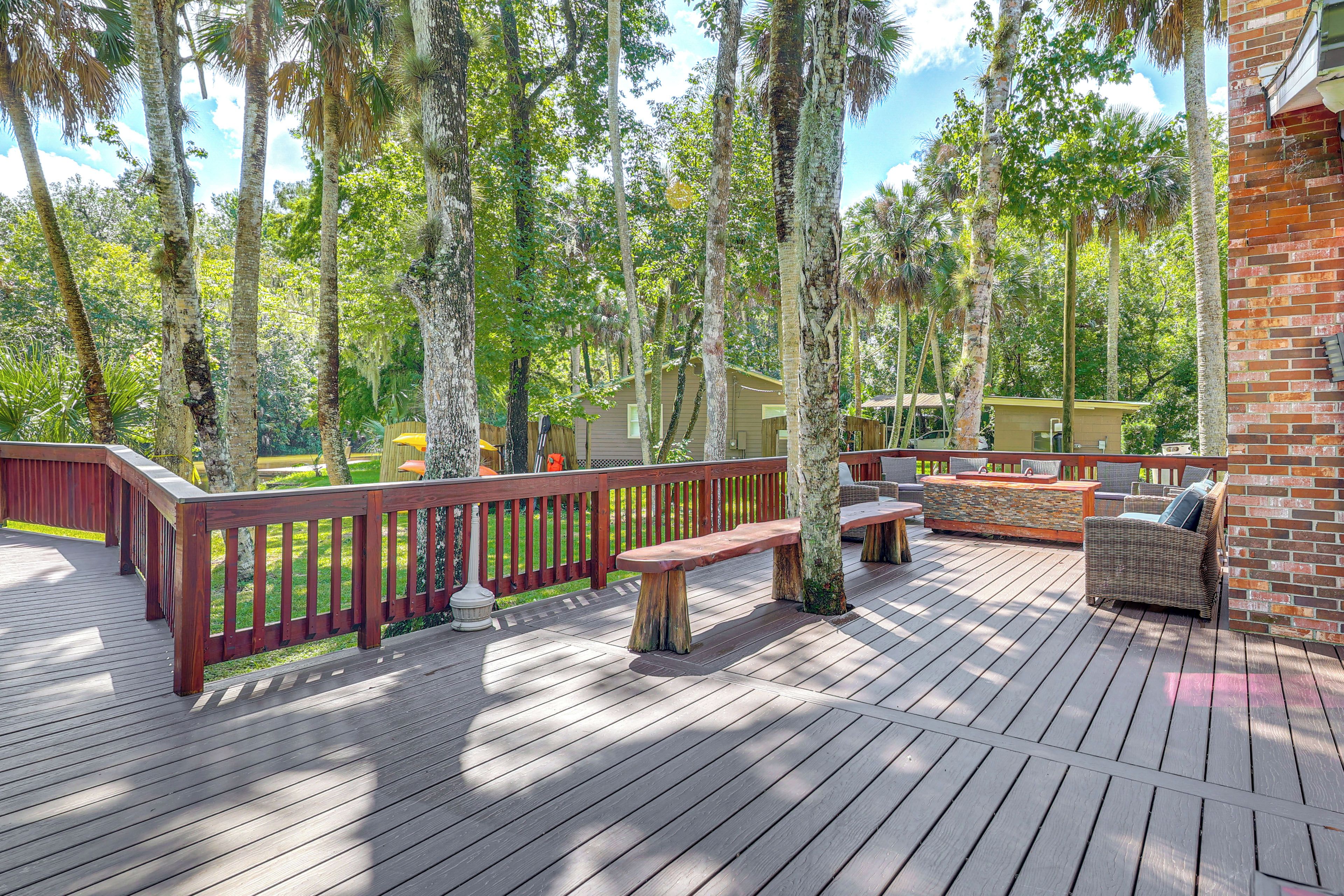 An outdoor deck with wooden railings, a seating area with a bench and a table, surrounded by trees.
