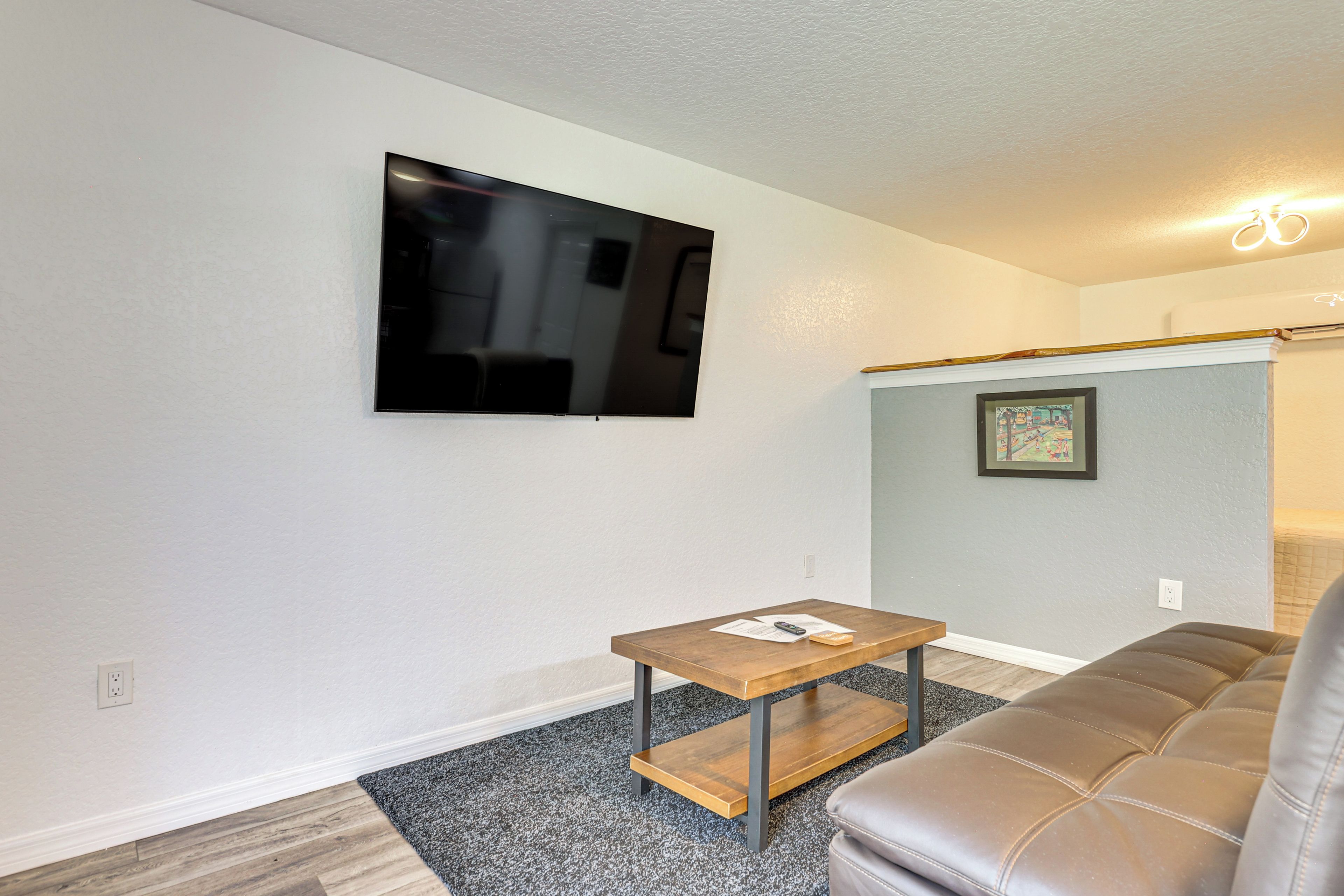 Una sala de estar con un televisor de pantalla plana montado en la pared, una pequeña mesa de madera, un sofá de cuero marrón y un cuadro enmarcado en la pared.