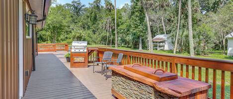 A wooden deck with a barbecue area, outdoor seating, and a view of a forested area.