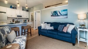 A modern living room with a blue sofa, a patterned armchair, and a kitchen area with white cabinets and pendant lighting.
