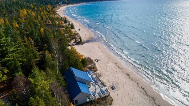An aerial view of a beach house with a blue roof, surrounded by dense forest, overlooking a sandy beach and calm sea. - Harbor Cove Condo | Ski • Beach • Explore (Harbor Springs)