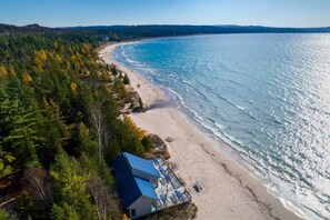 An aerial view of a beach house with a blue roof, surrounded by dense forest, overlooking a sandy beach and calm sea. - Harbor Cove Condo | Ski • Beach • Explore (Harbor Springs)