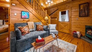 A log cabin interior with a staircase, a gray sofa with patterned pillows, a wooden coffee table, and a fireplace. - *Starlink Wifi* Peaceful Log Cabin Near National Park & Dollywood (Sevierville)