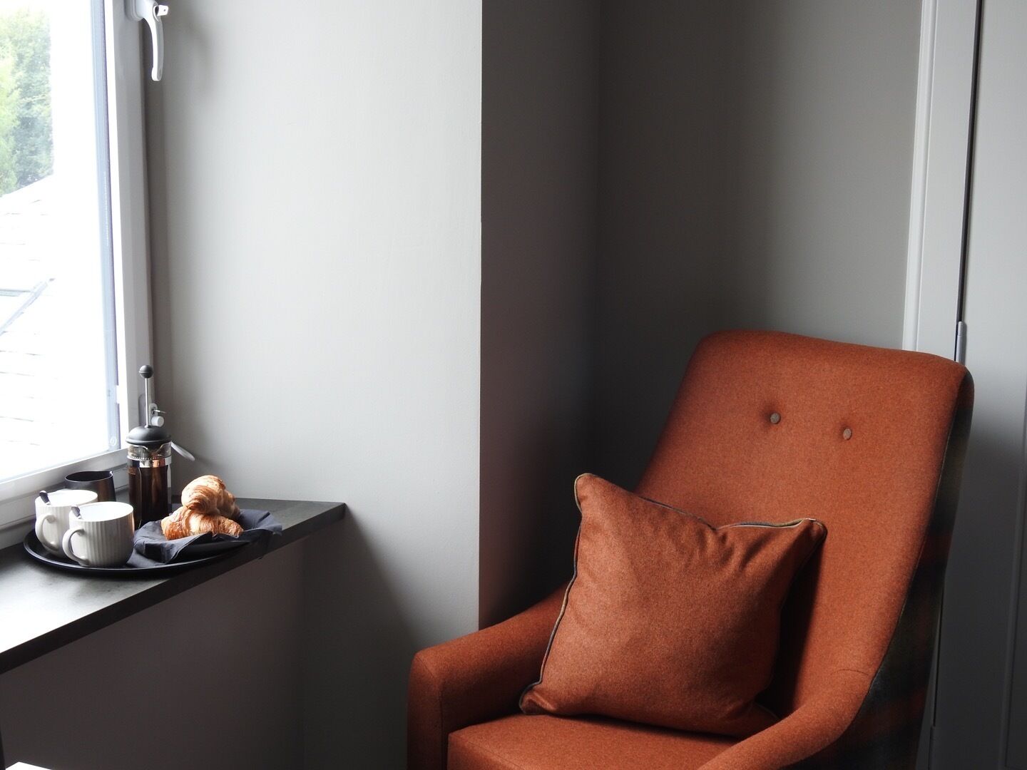 A corner room with a window, a small table with a cup, a mug, and a croissant, and an orange armchair with a pillow.