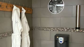 Un baño con un reloj de pared, un perchero con batas blancas y un secador de manos.