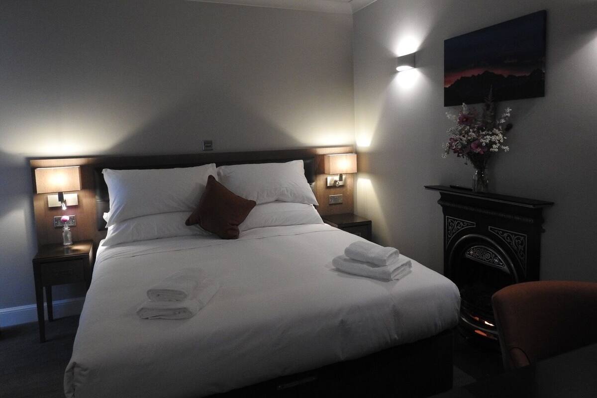 A neatly made bed with white linens and a brown pillow, two bedside lamps, a nightstand, a vase with flowers, and a painting on the wall.