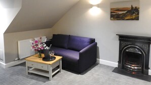 A cozy room with a purple sofa, a small wooden table with a vase of flowers, and a fireplace. - House Of Juniper - The Manse (Strath)