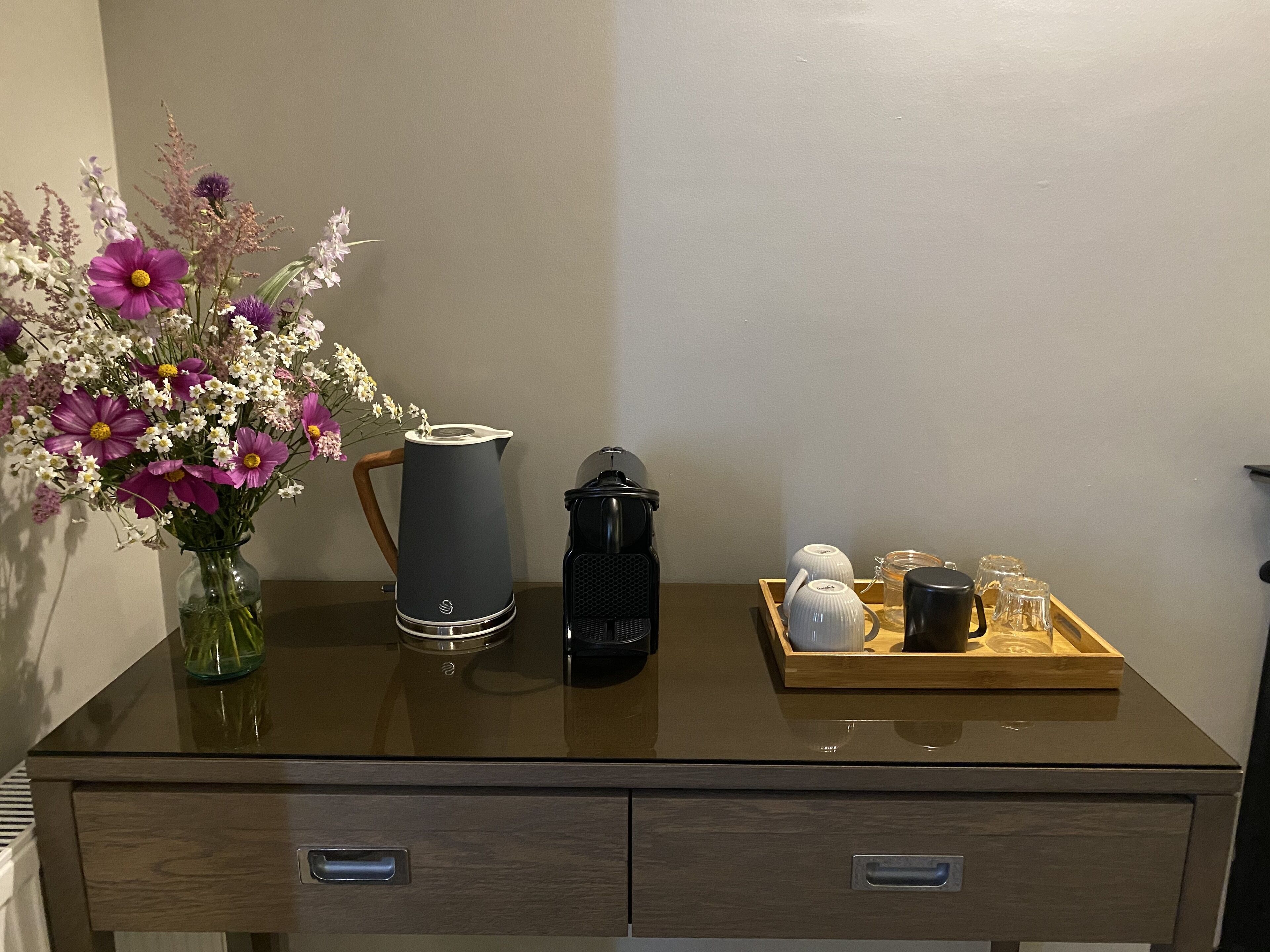 Un escritorio de madera con una cafetera, un florero con flores y una bandeja con tazas y una tetera.