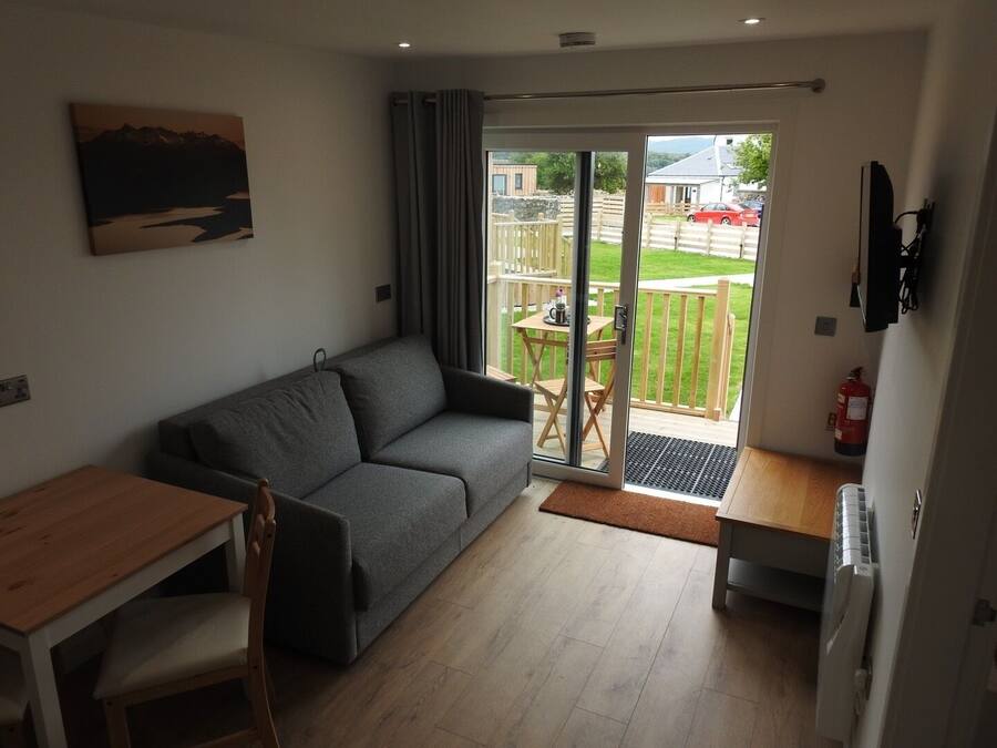 A modern living room with a grey sofa, wooden dining table, and chairs. There is a sliding glass door leading to a balcony with a wooden deck and a fire extinguisher mounted on the wall.