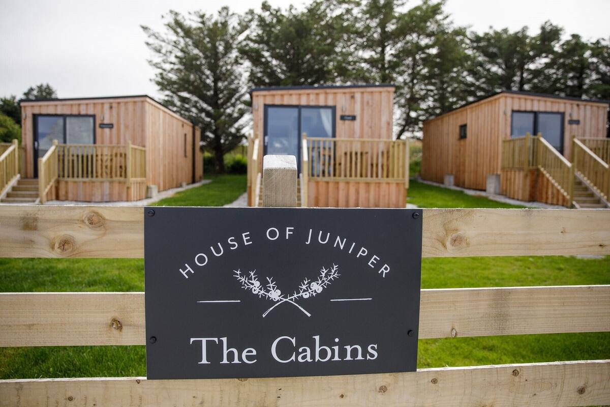 Un letrero que dice 'House of Juniper The Cabins' con cabañas de madera al fondo.