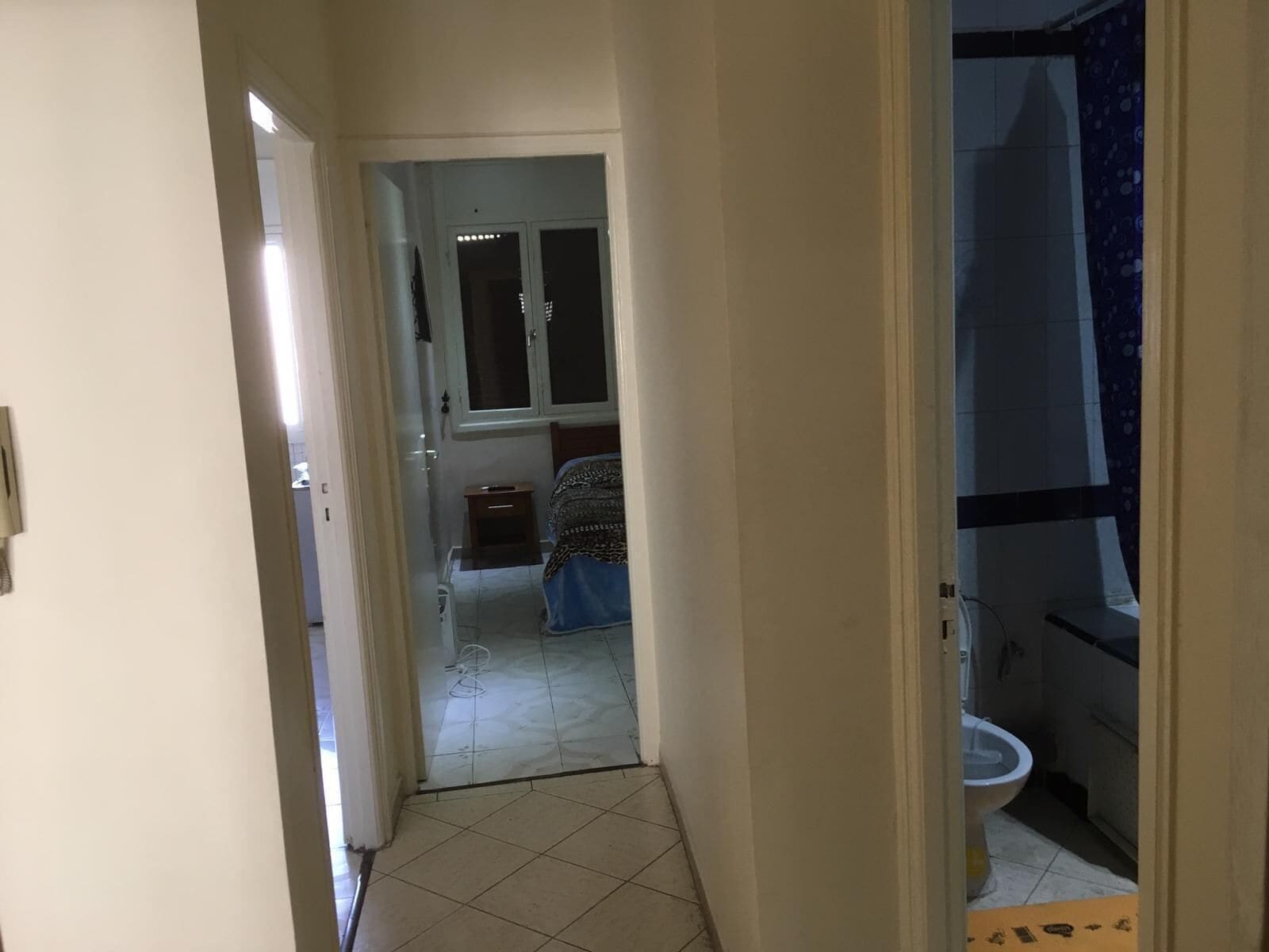Un pasillo estrecho que da a un dormitorio con cama y mesita de noche. A la derecha se ve un baño con inodoro y ducha.