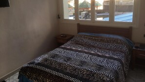 A bedroom with a bed, a nightstand, a window with a view of buildings, and a wall-mounted TV.