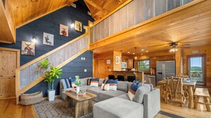 A cozy cabin interior with a living area featuring a gray sectional sofa, a wooden dining table, and a kitchen with a refrigerator and wooden cabinets. - Smoky/ Mountain views/ King bed/ bunk bed/en suite bath/Hot tub/Sauna/Pet/value (Sevierville)