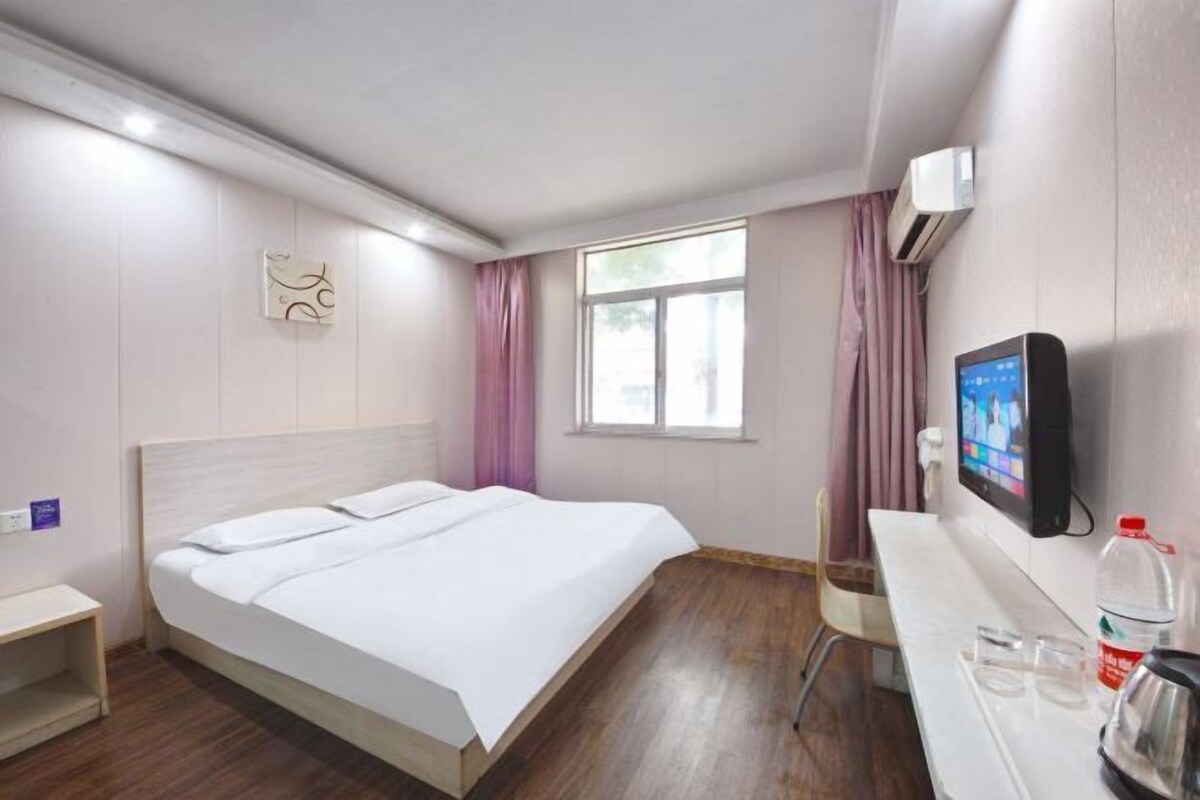 A hotel room with a bed, a desk, a chair, a TV, and an air conditioning unit.