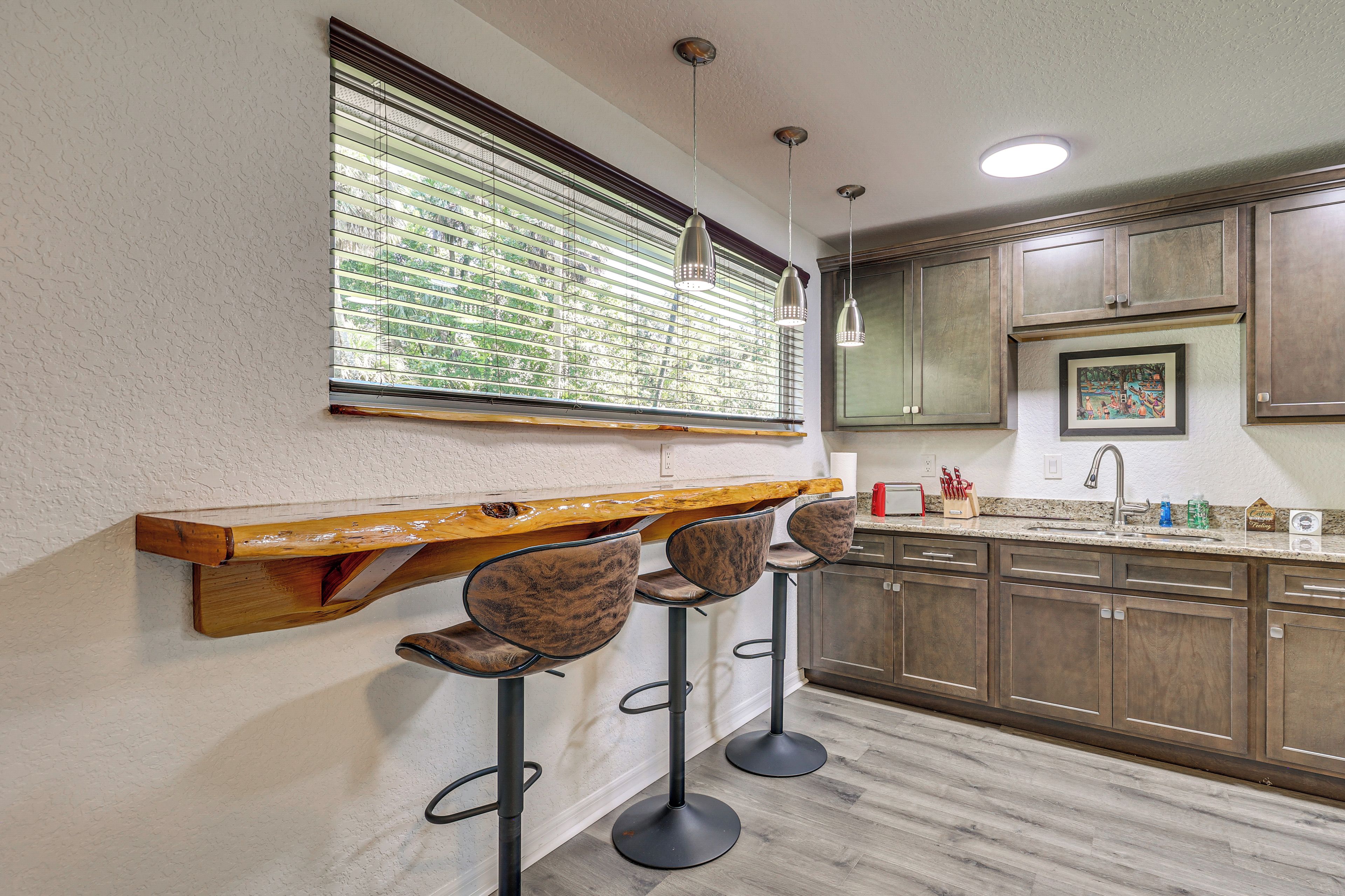 A kitchen with wooden cabinets, a bar area with stools, and a window with blinds.