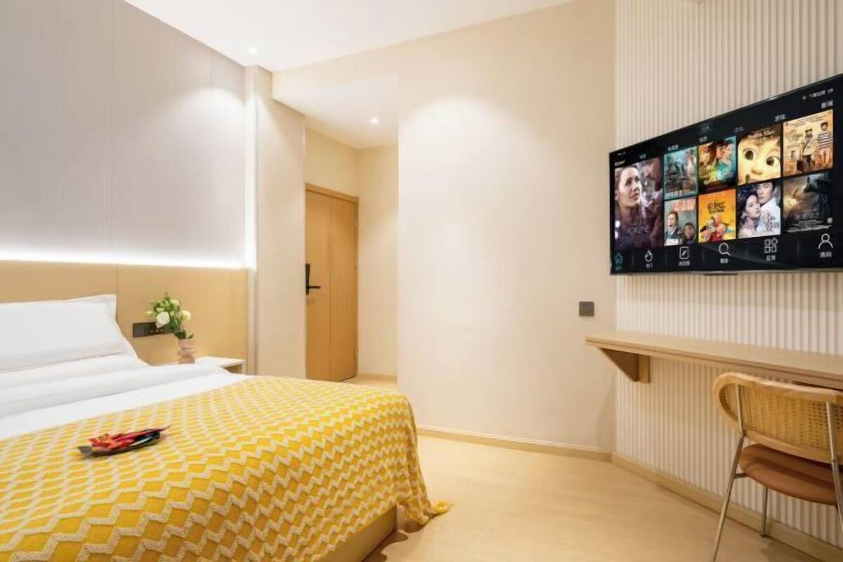A hotel room with a bed, a TV mounted on the wall displaying movie posters, a small table, and a chair.