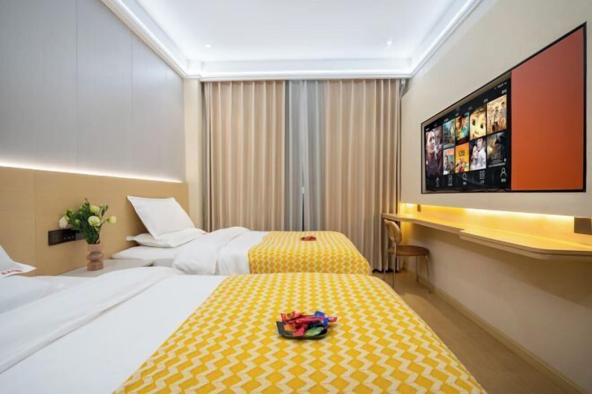 Habitación de hotel con dos camas, un ventanal grande con cortinas, un televisor de pantalla plana en la pared y una mesita con una silla.