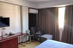 A hotel room with a bed, a desk with a flat-screen TV, a chair, a small table with a coffee cup, and an air conditioning unit.