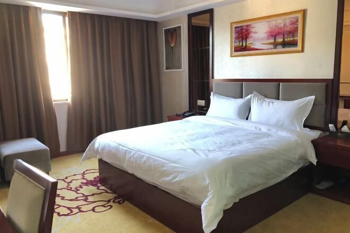 A hotel room with a large bed, a bedside table, a chair, a desk, and a painting on the wall.