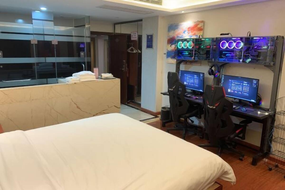Quarto de hotel com cama, escrivaninha com dois monitores e um computador com iluminação neon.