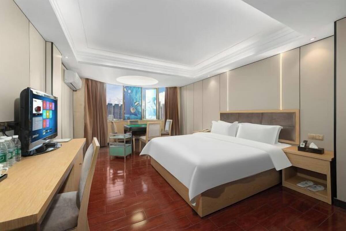 Quarto de hotel com uma cama grande, uma mesa de trabalho com um computador, uma televisão e uma janela com vista para a cidade.