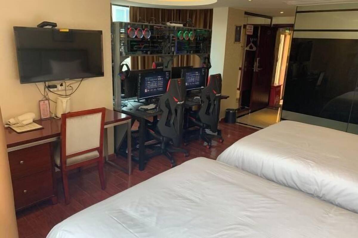 Quarto de hotel com uma mesa, dois monitores e uma cama.