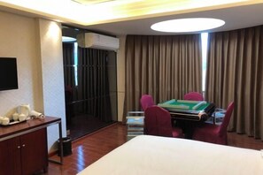 A hotel room with a bed, a desk with a computer, a chair, a table with a green tablecloth, a TV on the wall, and a window with curtains.