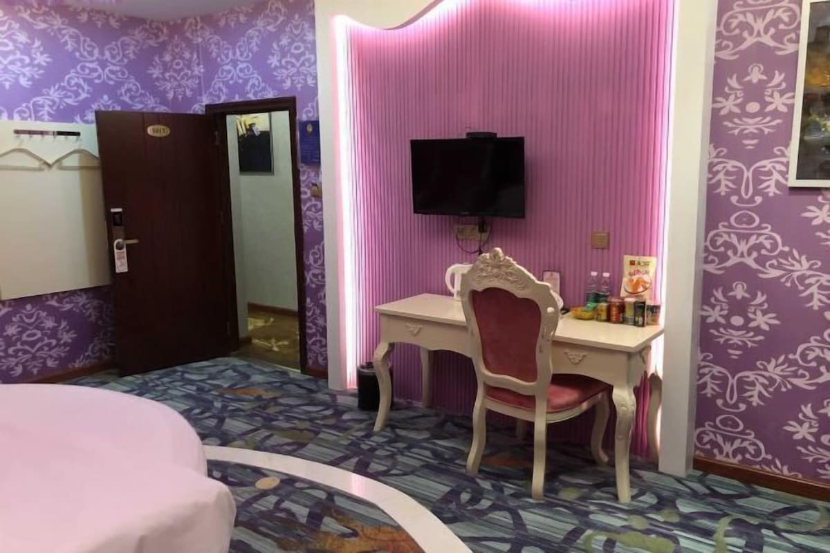 A hotel room with a bed, a desk with a chair, a television mounted on the wall, and patterned wallpaper.