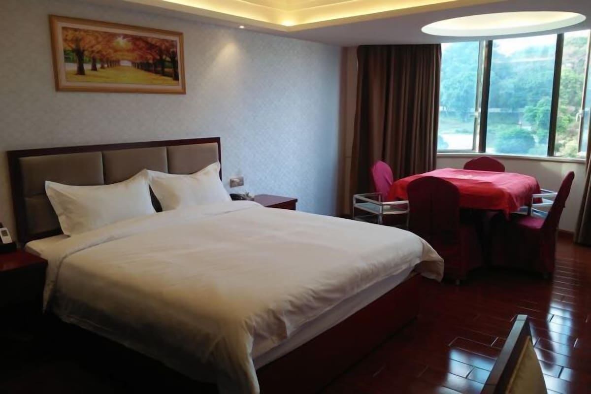 A hotel room with a large bed, a small table with chairs, a painting on the wall, and a window with a view of trees.
