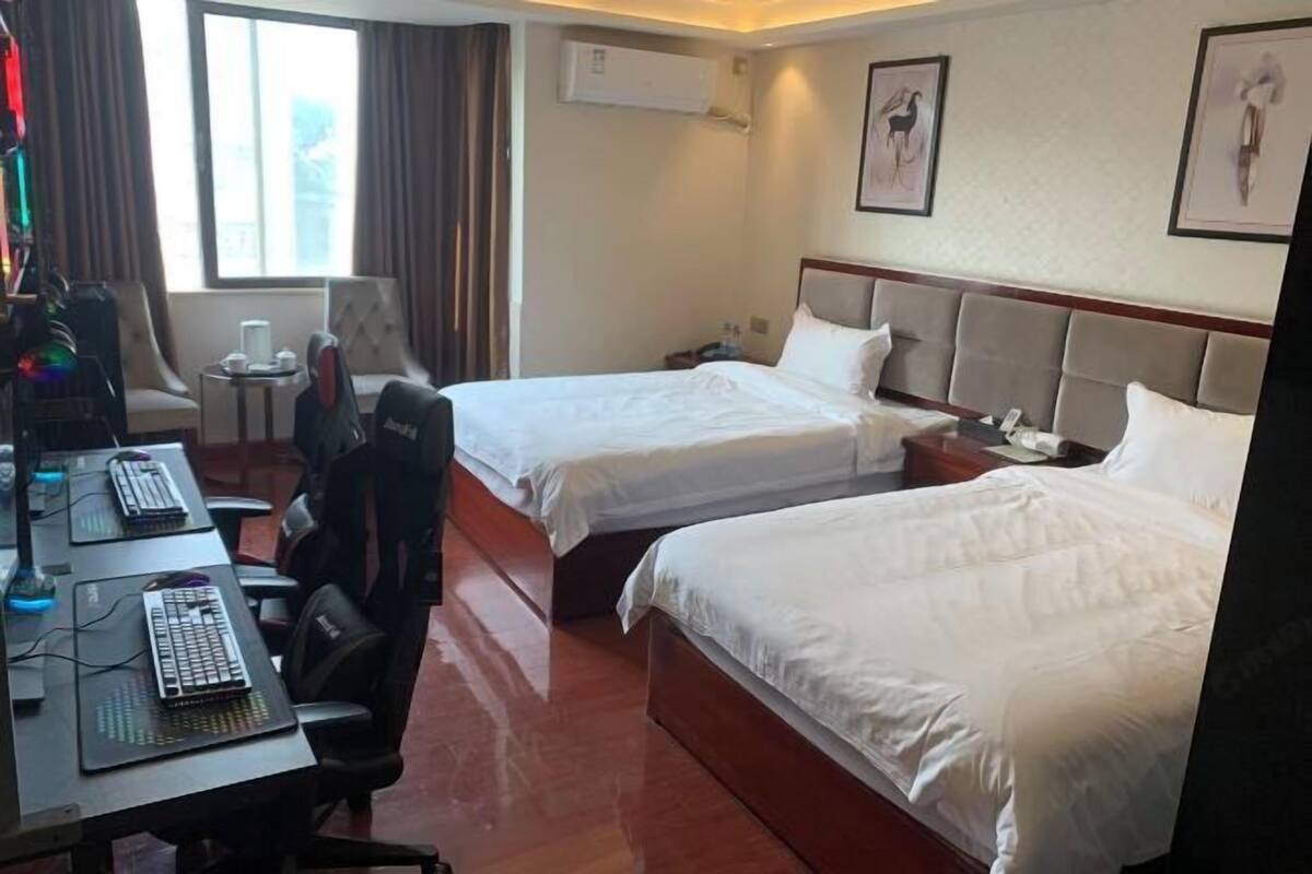 Quarto de hotel com duas camas, uma mesa com computador e teclado, uma cadeira, uma mesinha com abajur e quadros nas paredes.