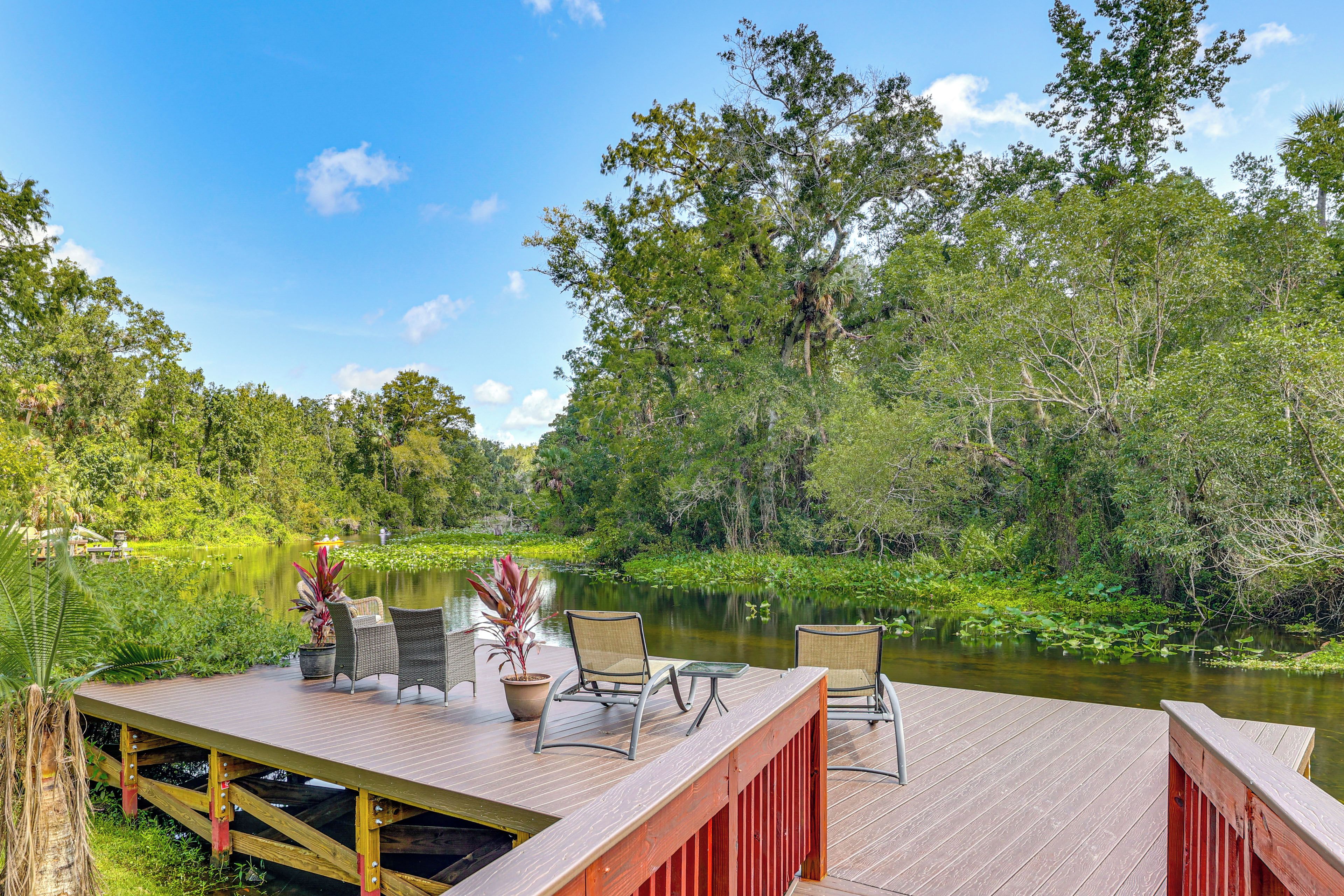 Une terrasse en bois, agréée de mobilier d’extérieur, qui offre une vue sur une rivière paisible, le tout entouré d’une végétation luxuriante et d’arbres.