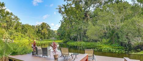 Une terrasse en bois, agréée de mobilier d’extérieur, qui offre une vue sur une rivière paisible, le tout entouré d’une végétation luxuriante et d’arbres.