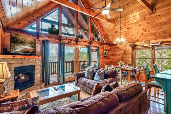 Un intérieur de chalet chaleureux avec une cheminée, un canapé en cuir, un coin repas et de grandes fenêtres offrant une vue sur l’extérieur.