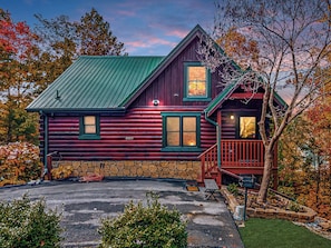 A log cabin with a stone foundation, a green metal roof, and a wooden deck.