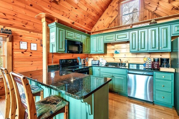 Una cocina con gabinetes verdes, encimera de granito, lavavajillas de acero inoxidable y suelo de madera.