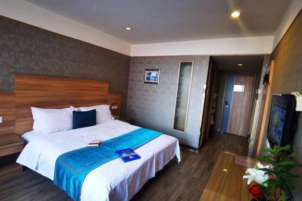 A hotel room with a large bed, wooden headboard, a small bedside table, a nightstand, a TV, and a potted plant.