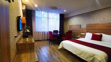 A hotel room with a wooden floor, a bed with white and red bedding, a desk with a chair, a TV on the wall, and a window with curtains.