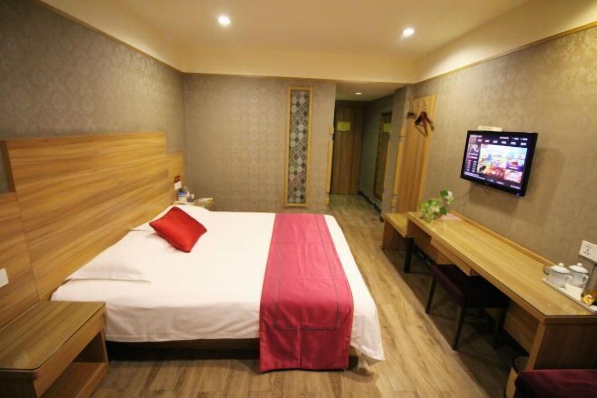 A hotel room with a wooden headboard bed, a desk with a chair, a television mounted on the wall, and a small table with a plant.