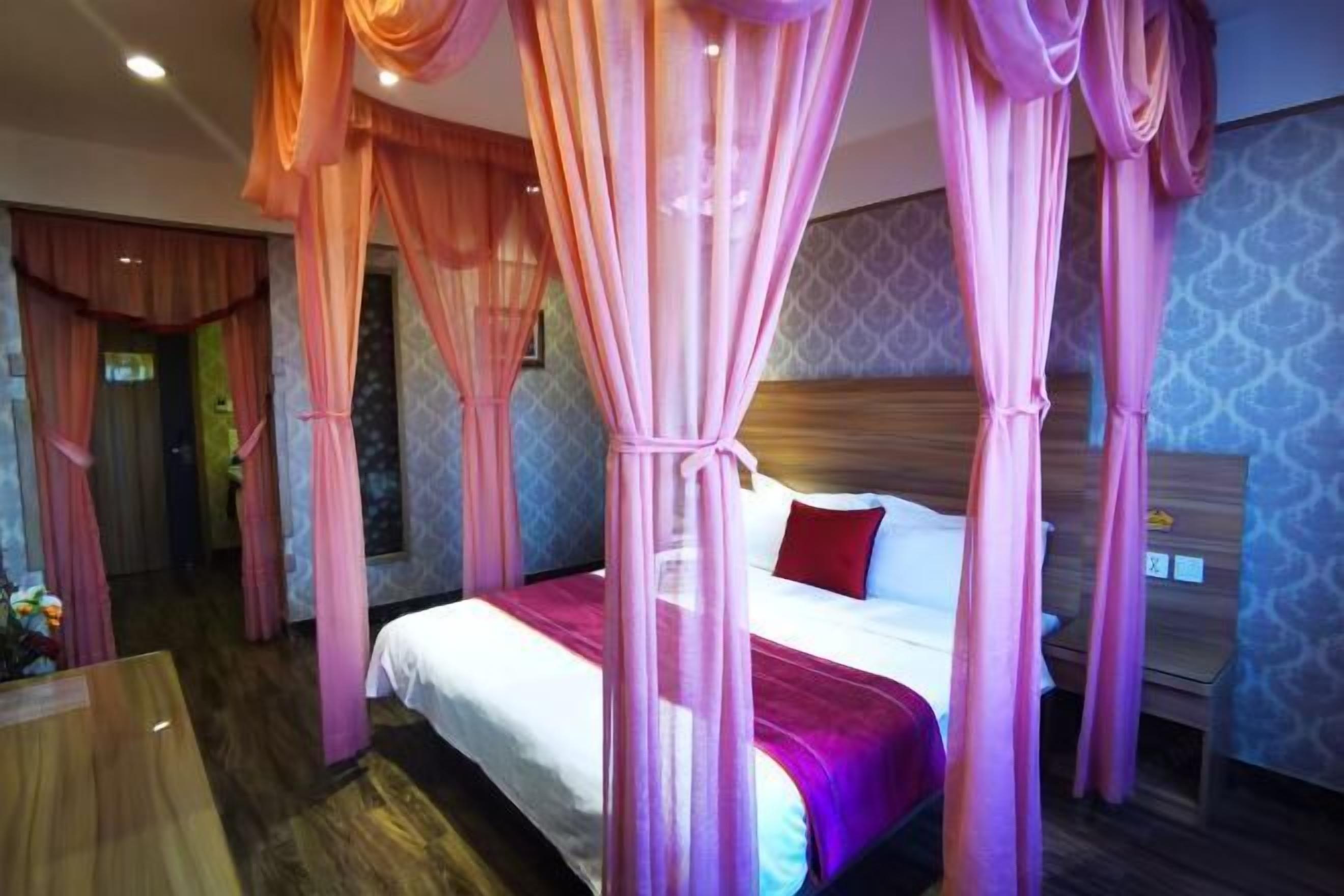 Quarto de hotel com uma cama, cabeceira de madeira e cortinas cor-de-rosa.