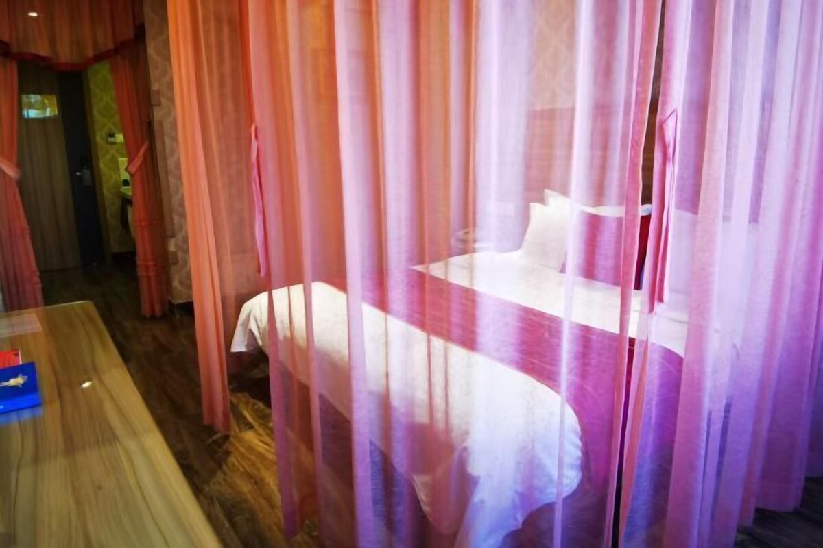 A room with a bed covered by sheer curtains, a wooden table, and a visible bathroom area.