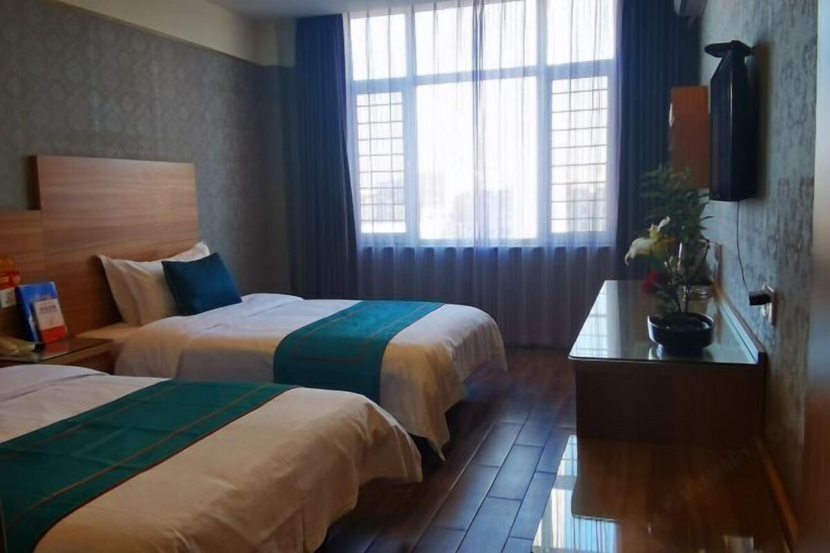 A hotel room with two beds, a wooden headboard, a bedside table with a plant, and a large window with curtains.
